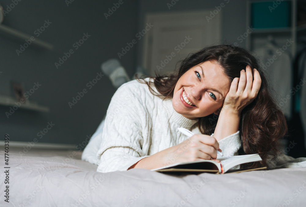 Young adult happy brunette woman forty years plus size body positive in white knitted sweater writes notes in planner on bed at the home Young adult happy brunette woman forty years plus size body positive in white knitted sweater writes notes in planner on bed at the home