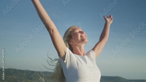 Blond female breathing in fresh mountain air on summit smiling pure happiness raising arms in air