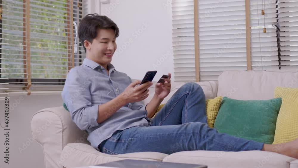 Asian man makes a payment via the internet. On the sofa at home, a shopper shops online using a credit card and a smartphone. The concept of internet shopping. Asian man makes a payment via the internet. On the sofa at home, a shopper shops online using a credit card and a smartphone. The concept of internet shopping.
