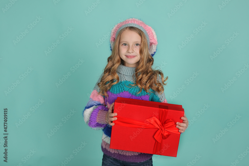 a beautiful happy blonde girl in a colored knitted sweater and hat in her hands a red gift box on a blue background a beautiful happy blonde girl in a colored knitted sweater and hat in her hands a red gift box on a blue background