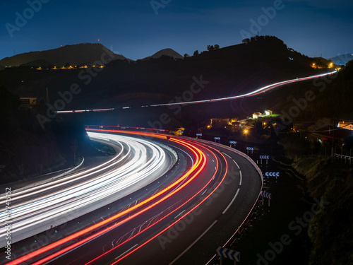 Trazos de luz dejados por el tráfico al circular por la noche por una autopista a su paso por un desfiladero, con las luces de una ciudad al fondo. photo
