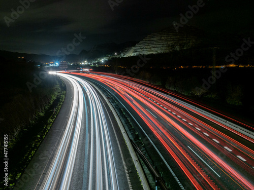 Trazos de luz dejados por el tráfico al circular por la noche por una autopista a su paso por un desfiladero, con las luces de una ciudad al fondo. photo