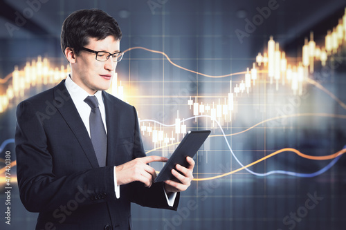 Attractive young european businessman using tablet with abstract glowing candlestick forex hologram in blurry office interior. Trade and finance concept. Double exposure.