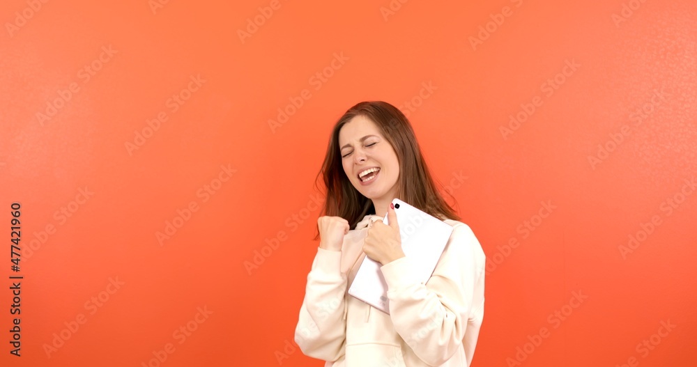 A young female student in a hoodie makes notes on a tablet on an orange background. A woman writes with a pen on a new gadget. A young female student in a hoodie makes notes on a tablet on an orange background. A woman writes with a pen on a new gadget.