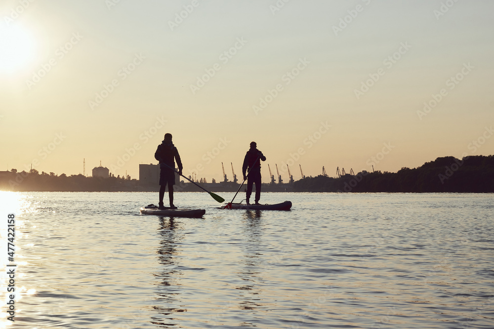 Silhouettes of two boys rowing on supboards (SUP) at Danube river at sunset