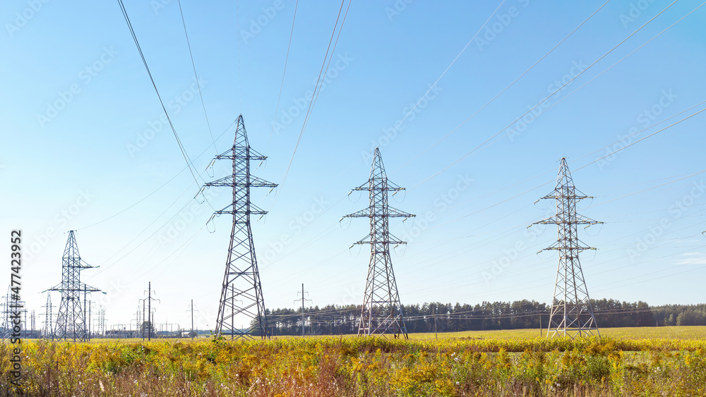 High voltage towers with blue sky background. Overhead power lines on the background of forest and green field. Electricity transmission through power lines. Green energy.