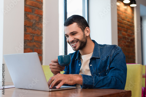 cheerful man typing on laptop while drinking coffee in restaurant