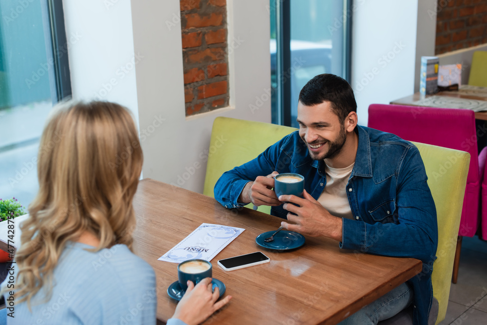 Fototapeta premium cheerful man drinking coffee with girlfriend near smartphone with blank screen and menu on table
