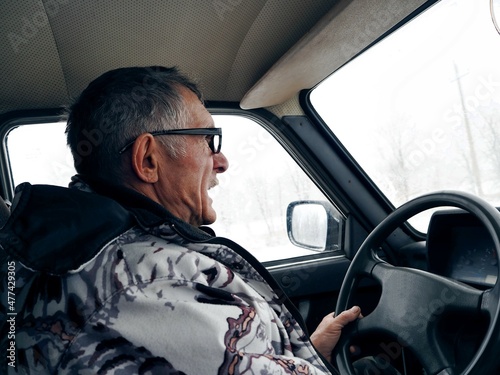An elderly man in a winter jacket drives a Lada car in winter on a fishing trip. He is nervous and screams, as it is winter and snow behind the glass of the windows. © Татьяна Пинкасевич