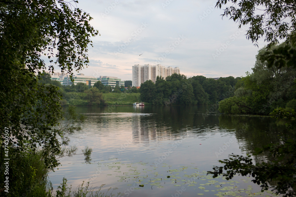 Beautiful nature with pond lake, green trees foliage, grass, buildings and  clouds in the background. Afternoon panorama landscape at Pokrovskoe Streshnevo urban forest park, Moscow, Russia