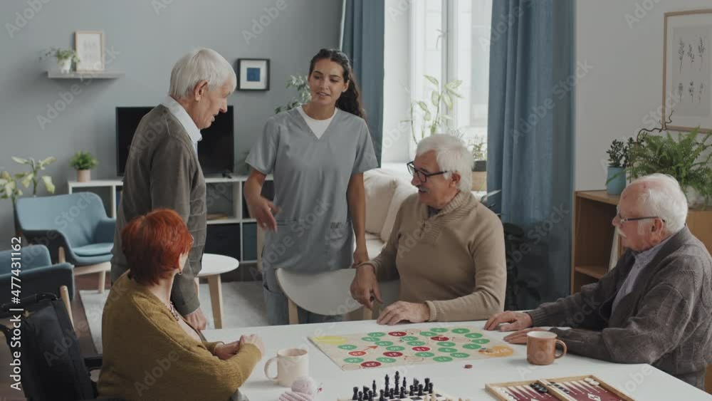 Medium shot of kind female nurse introducing Caucasian senior man to group of elderly people sitting around table in nursing home playing board games