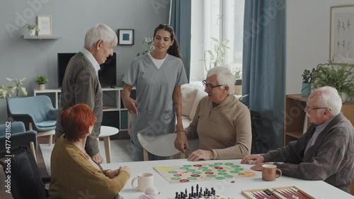 Medium shot of kind female nurse introducing Caucasian senior man to group of elderly people sitting around table in nursing home playing board games photo