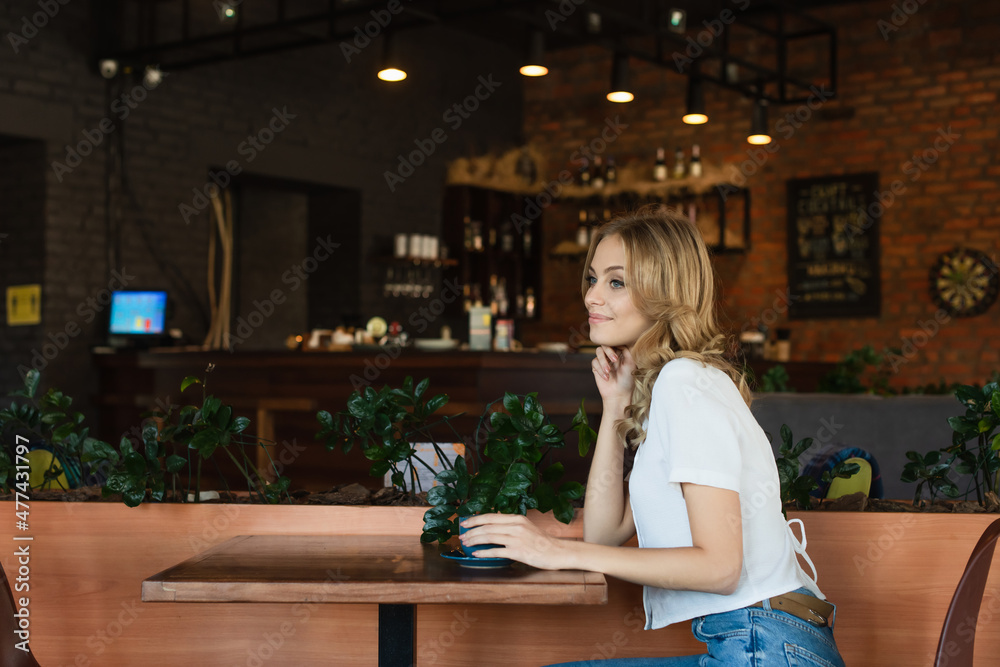 pretty and happy woman looking away while sitting in cafe near coffee cup pretty and happy woman looking away while sitting in cafe near coffee cup
