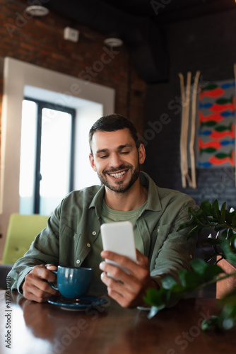 happy man looking at mobile phone while holding coffee cup in restaurant