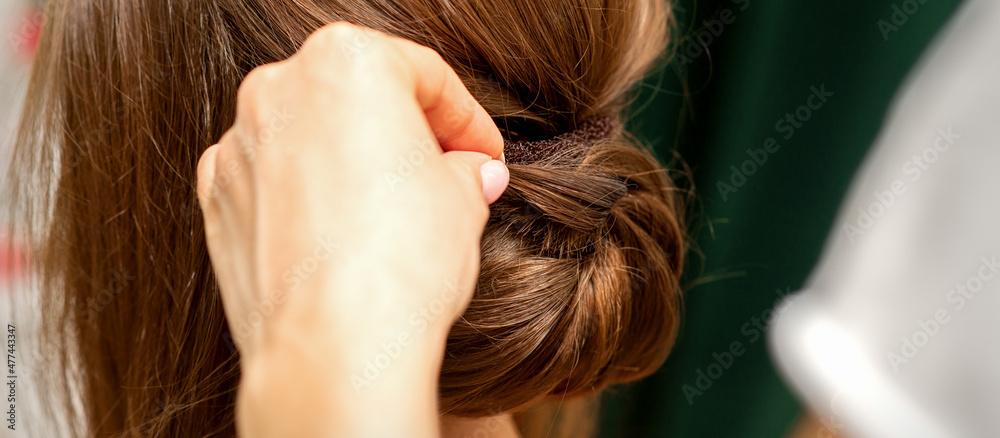 Fototapeta premium Hands of hairdresser making french twist hairstyle of an unrecognizable young brunette woman in a beauty salon, back view, close up