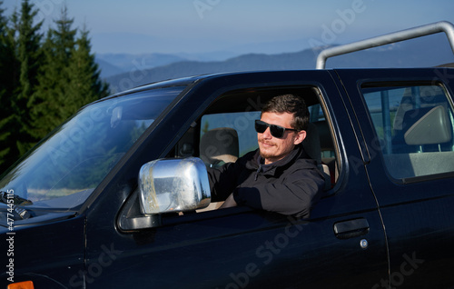 Assured man with smile on his face in black sunglasses behind the wheel of SUV, leaning his elbow on side door. Green trees and mountain hills on the background.