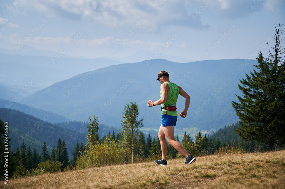 Tall sportsman running trail downhill during active holiday in the mountains. Jogging on mountain meadows against backdrop of panorama mountains.
