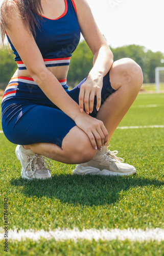 A young girl is sitting on a green lawn. Sportswear and shoes. Sports and fitness classes. Football field.