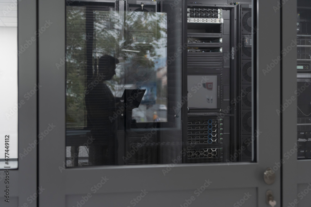 Software Engineer Working on a Laptop Computer in a Modern Server room. Monitoring Room Big Data Scientist in reflection of the entrance door. Software Engineer Working on a Laptop Computer in a Modern Server room. Monitoring Room Big Data Scientist in reflection of the entrance door.
