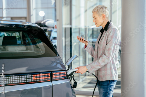 Eco electric car concept. Blond female waiting electric car to charge. Business woman with the charging cable near the electic vehice, hybrid cars. Concept of buying electric vehicle.