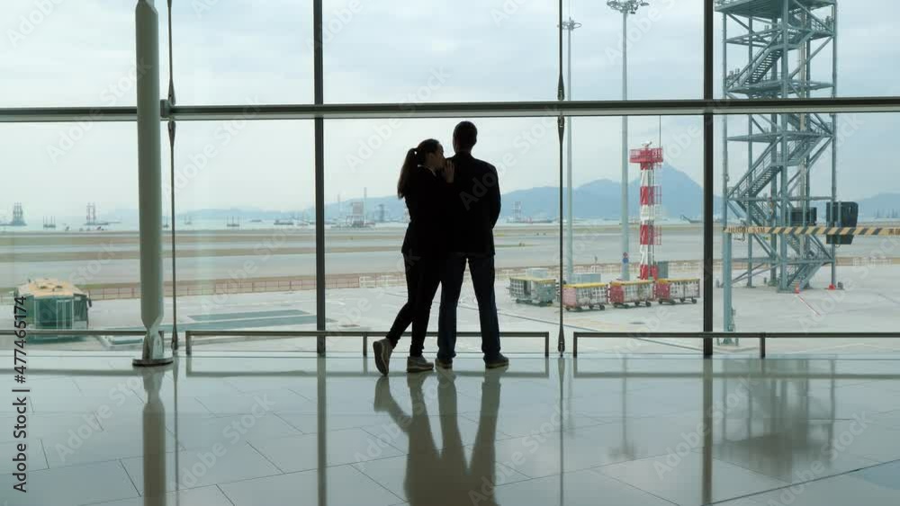 Two people stand against glass wall of terminal, woman embrace and lean on boyfriend shoulder. Man look out, silhouetted shot of travelers couple, pensive mood