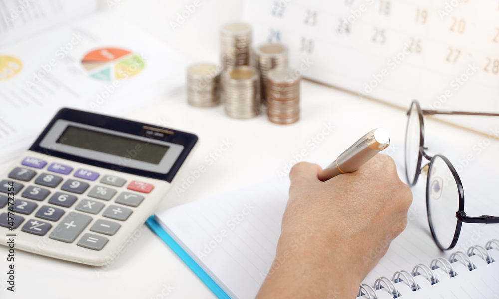 female hand writing a notebook, calculator and Money coins stack and pen for finance concept. female hand writing a notebook, calculator and Money coins stack and pen for finance concept.