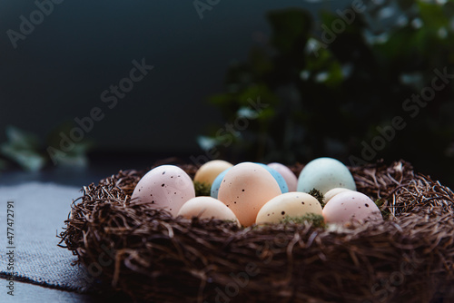 A wicker nest with moss and colored Easter eggs on gray linen fabric napkin and few green leaves on dark background. Dark mood style. Happy Easter holiday. Selective focus, copy space