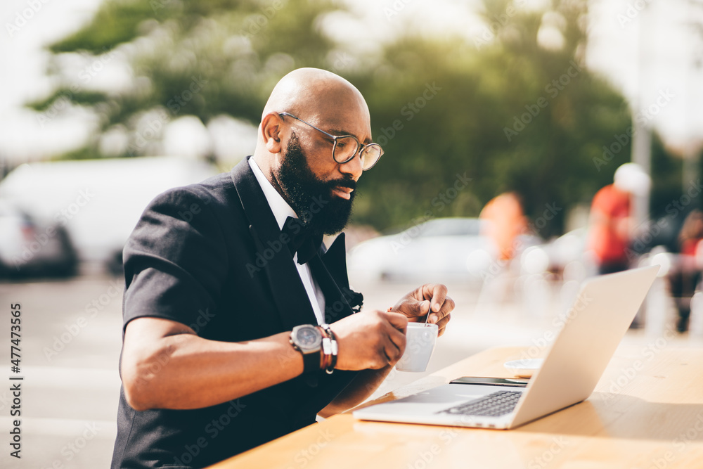 Selective focus on a stylish bearded gentleman sitting outdoors, using a notebook, and holding a cup. A black man in glasses at a cafe table, looking at his notebook screen while stirring his coffee - obrazy, fototapety, plakaty 