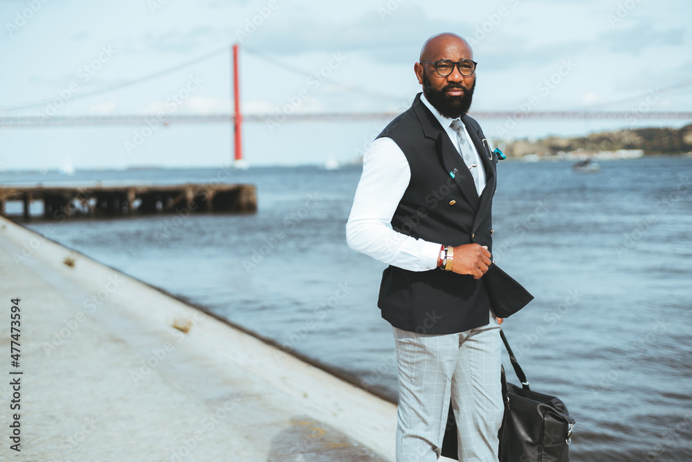 A businessman holding a messenger bag near a river, a red suspension bridge in a blurred background. A bald bearded black guy in a suit standing on the pier looking away, a copy space area on the left