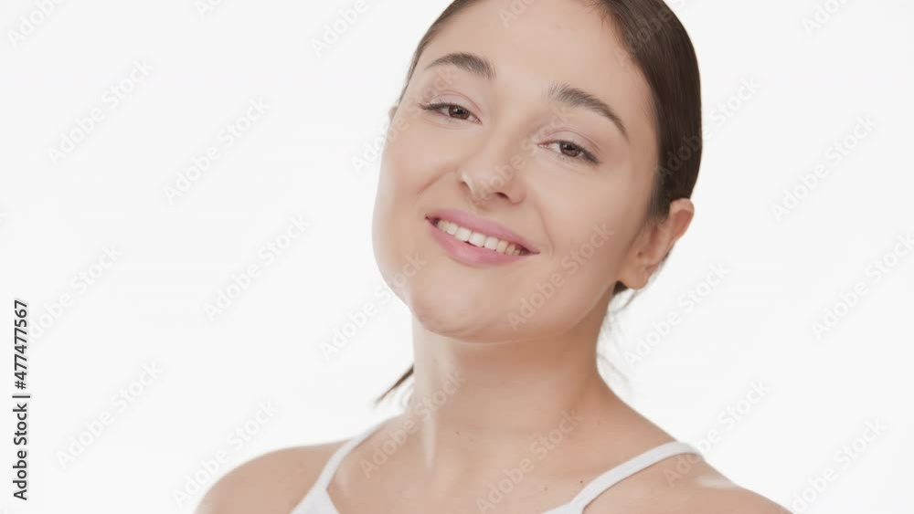 Horizontal close-up of attractive Caucasian young plus size woman with long brown hair in white top touches her cheek and looks at the camera smiling wide on white background | Skin nourishing concept Horizontal close-up of attractive Caucasian young plus size woman with long brown hair in white top touches her cheek and looks at the camera smiling wide on white background | Skin nourishing concept