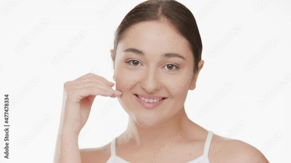 Horizontal close-up of gorgeous Caucasian young plus size brown-haired woman in white top touches her smooth face smiling wide for the camera on white background | Acne prevention concept Horizontal close-up of gorgeous Caucasian young plus size brown-haired woman in white top touches her smooth face smiling wide for the camera on white background | Acne prevention concept