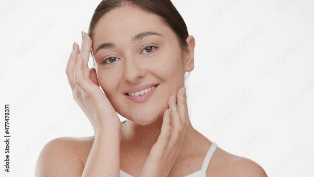 Beautiful Caucasian young plus size woman with long brown hair in white top strokes her face tilting her head from side to side and smiling wide on white background | Anti aging concept Beautiful Caucasian young plus size woman with long brown hair in white top strokes her face tilting her head from side to side and smiling wide on white background | Anti aging concept
