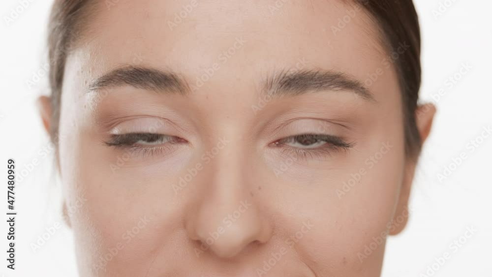 Horizontal extreme close-up of good-looking Caucasian young woman with dark hair blinks occasionally looking at the camera on white background | Droopy eyes removal concept