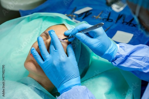 Woman patient is lying on the operating table. Doctor wearing medical gloves is cutting the skin above her eyebrow with a scalpel