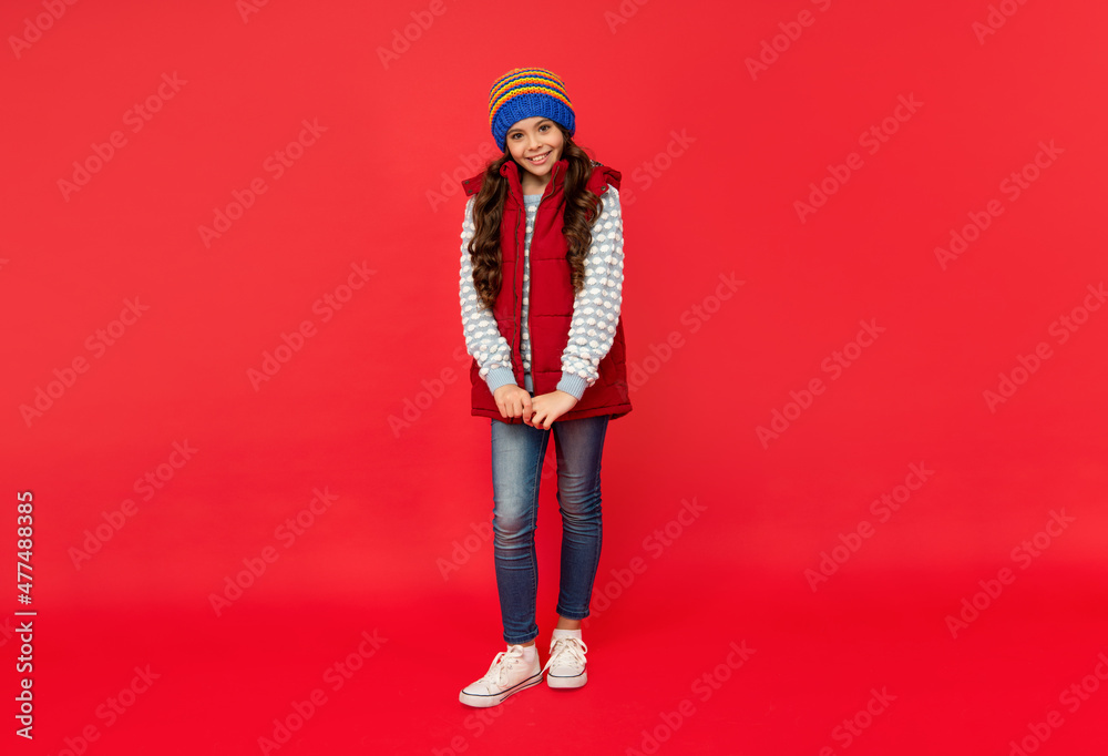 full length of happy teen girl in knitted winter hat and down vest on red background, winter outfit full length of happy teen girl in knitted winter hat and down vest on red background, winter outfit