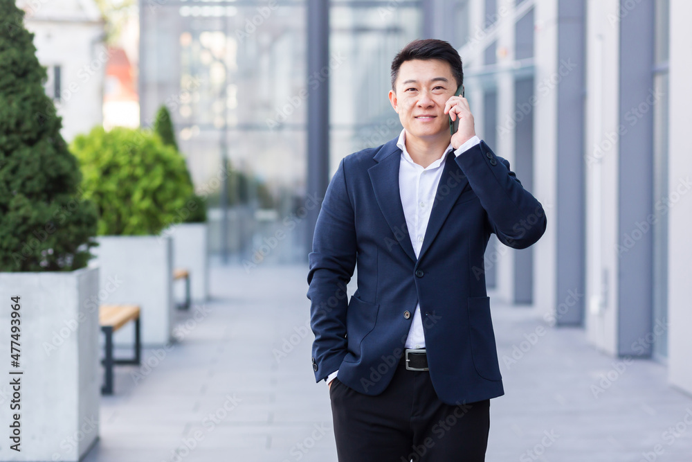 Successful asian businessman businessman talking on cell phone while walking near big modern office, cheerful smiling worker and looking at camera Successful asian businessman businessman talking on cell phone while walking near big modern office, cheerful smiling worker and looking at camera