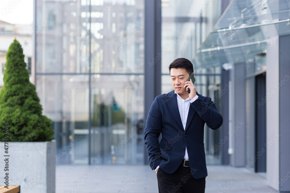 Successful Asian businessman businessman talking on a mobile phone while walking near the office of a large modern, serious and pensive employee Successful Asian businessman businessman talking on a mobile phone while walking near the office of a large modern, serious and pensive employee