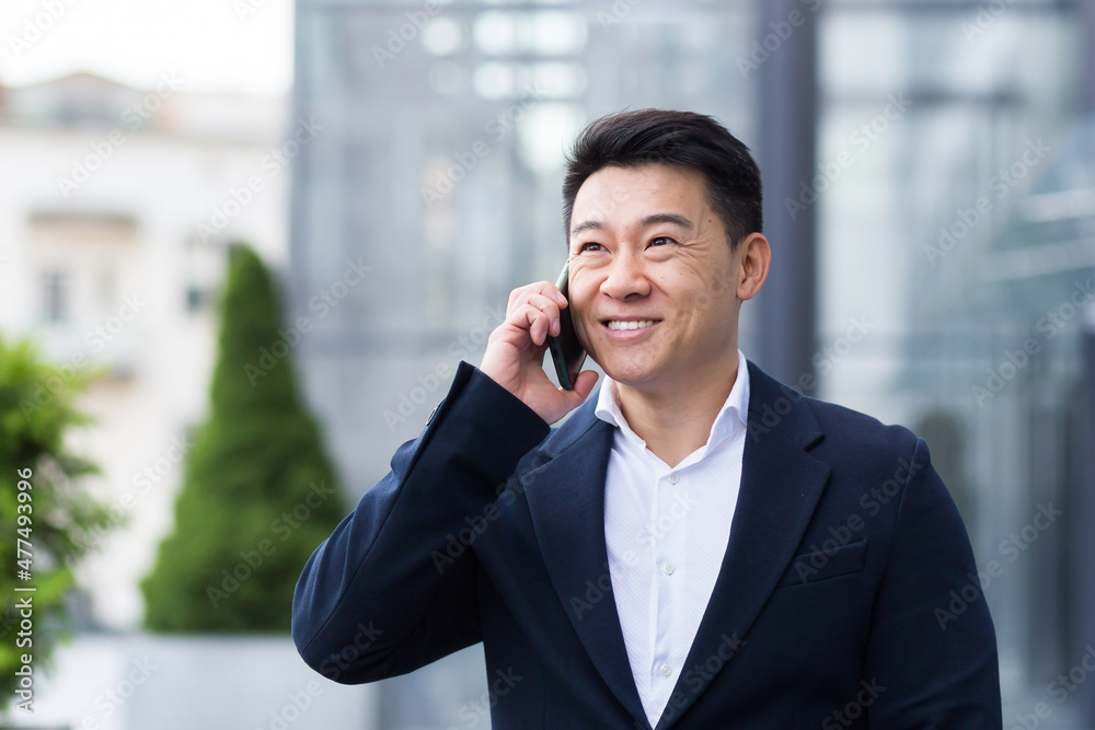 Successful asian businessman businessman talking on cell phone while walking near big modern office, cheerful smiling worker and looking at camera Successful asian businessman businessman talking on cell phone while walking near big modern office, cheerful smiling worker and looking at camera