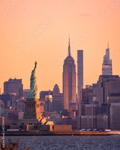 New York, NY - USA -Dec 26, 2021: Vertical early morning view of the Statue of Liberty in the New York Harbor, with the Empire State Building and the skyline of Manhattan behind her at sunrise.