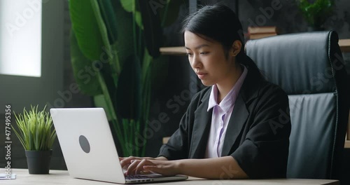 Asian businesswoman working on laptop computer at home office. Female manager typing on computer keyboard indoors. Portrait of serious business woman looking at laptop screen in modern office