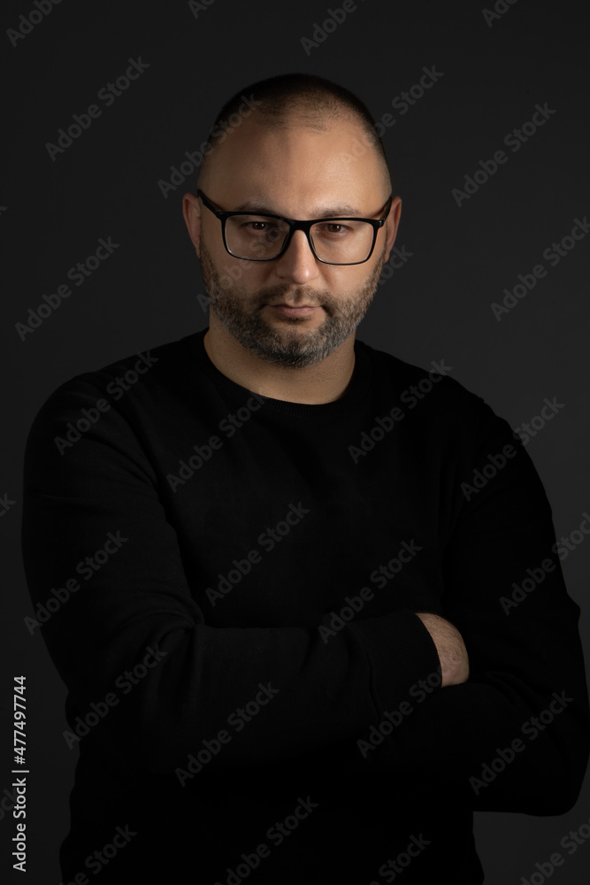 A young man with a short haircut, black-rimmed glasses and a black sweater crossed his arms over his chest and poses looking at the camera A young man with a short haircut, black-rimmed glasses and a black sweater crossed his arms over his chest and poses looking at the camera