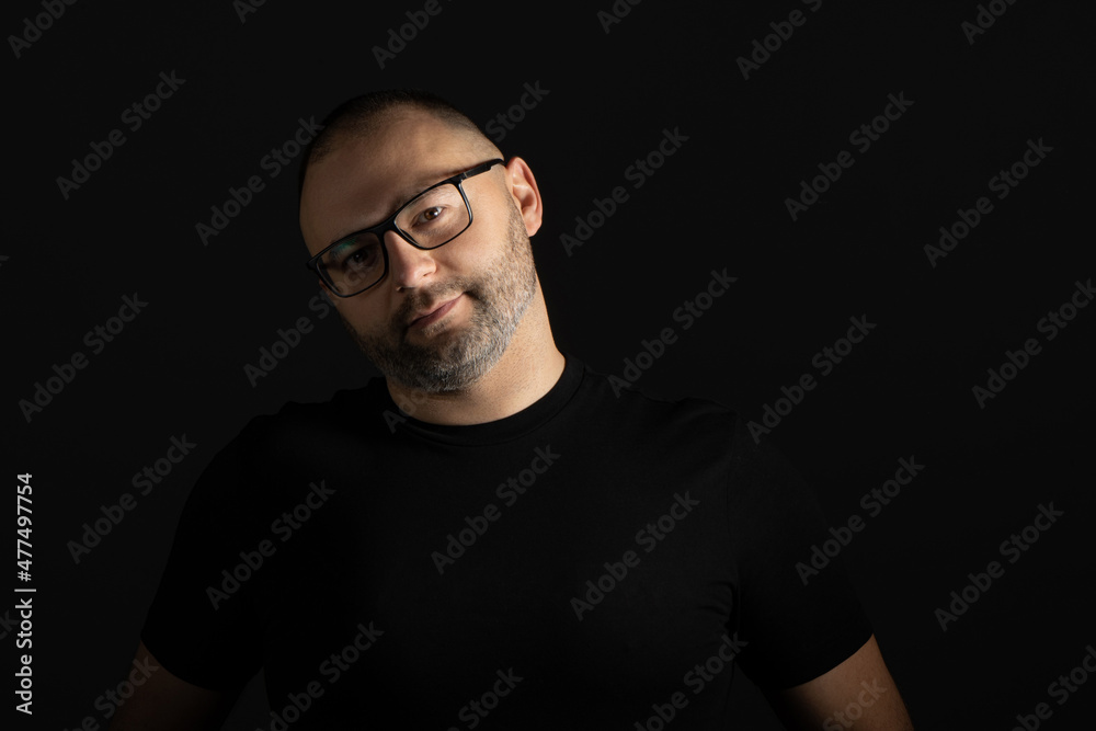 A young man in a black T-shirt with a short haircut and black-rimmed glasses poses against a dark background carefully and aggressively looking at the camera