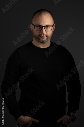A young man in black-rimmed glasses and a black sweater keeps his hands in his pockets posing for the camera