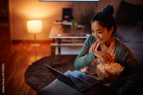 Canvas Print Happy Asian woman eats popcorn while watching movie on laptop at night at home
