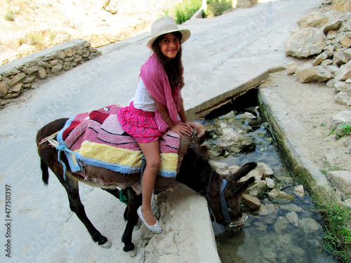 Cute young girl rides a donkey in a desert oasis. Donkey drinks water photo