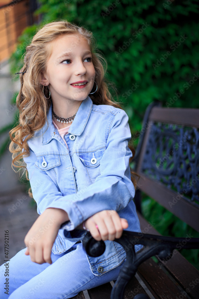 Portrait of a joyful blonde-haired blue-eyed girl sitting on a park bench. The girl is wearing a blue denim jacket Portrait of a joyful blonde-haired blue-eyed girl sitting on a park bench. The girl is wearing a blue denim jacket