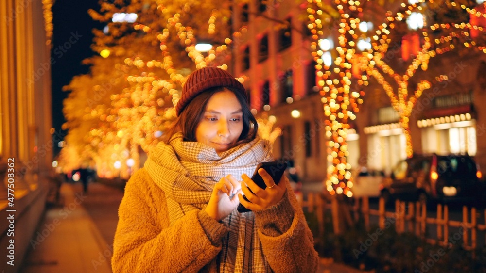 Young Woman Walking And Using Smartphone In The Evening Winter Street. Social Networking New Year Christmas Concept Photo.