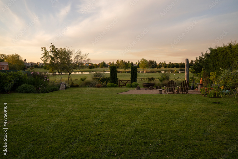 Real estate. End of day in the suburbs. View of the park with short green grass, at sunset. The golf course with an artificial pond in the background. 