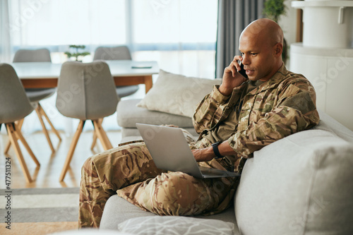 Black military man uses laptop while talking on cell phone at home.