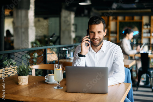 Smiling businessman talks on cell phone while using laptop in cafe.